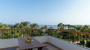 A balcony with a wooden table and a bowl of fruit, overlooking a coastal view with palm trees and the sea.