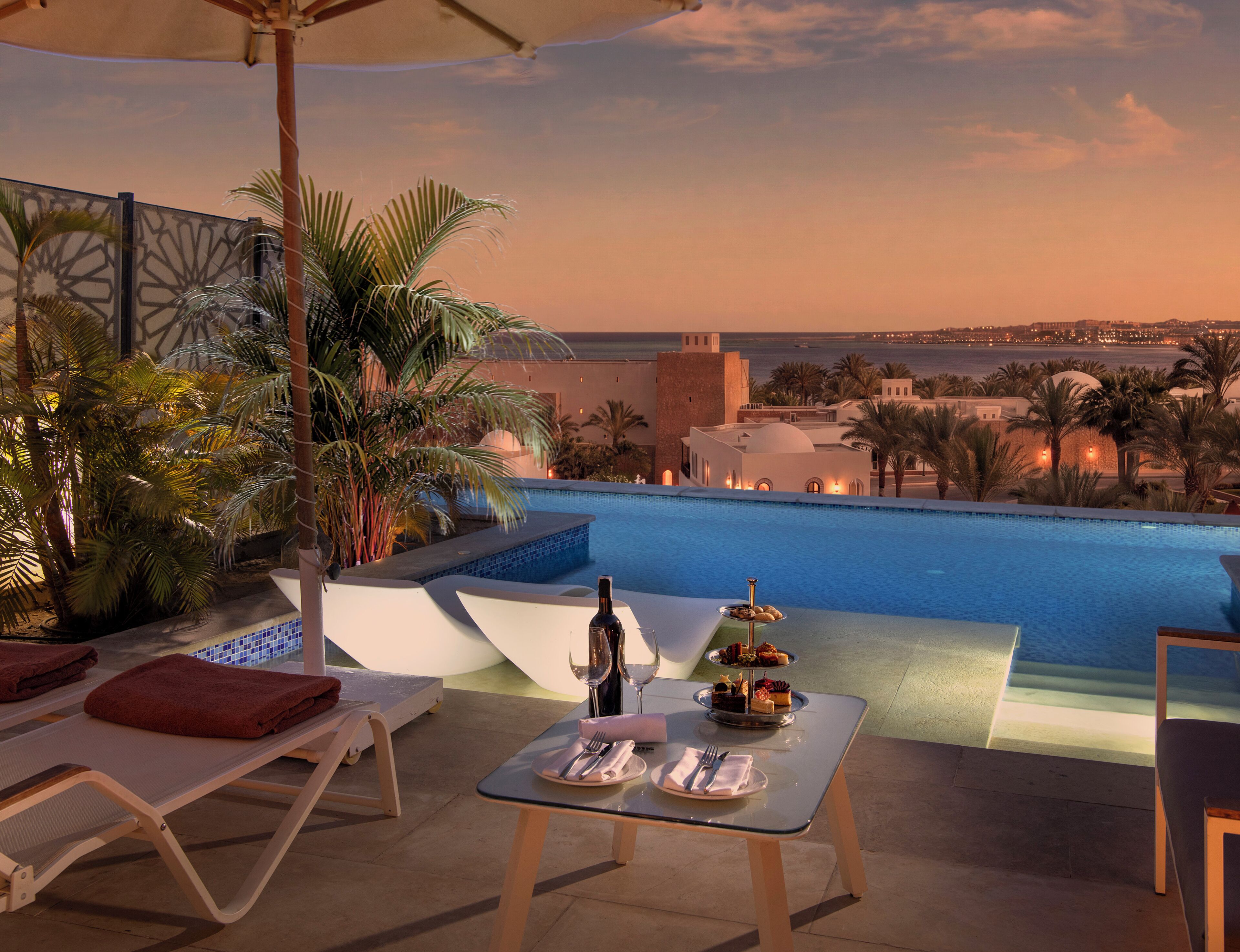 Un área de comedor junto a la piscina con vista al mar y una estructura que recuerda a un castillo al fondo.