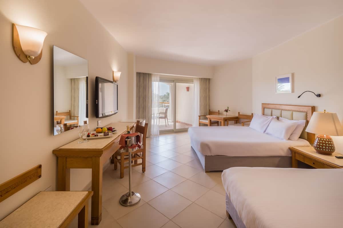 Ett hotellrum med två sängar, ett skrivbord med en stol, en TV och ett matbord med frukt och en flaska champagne.