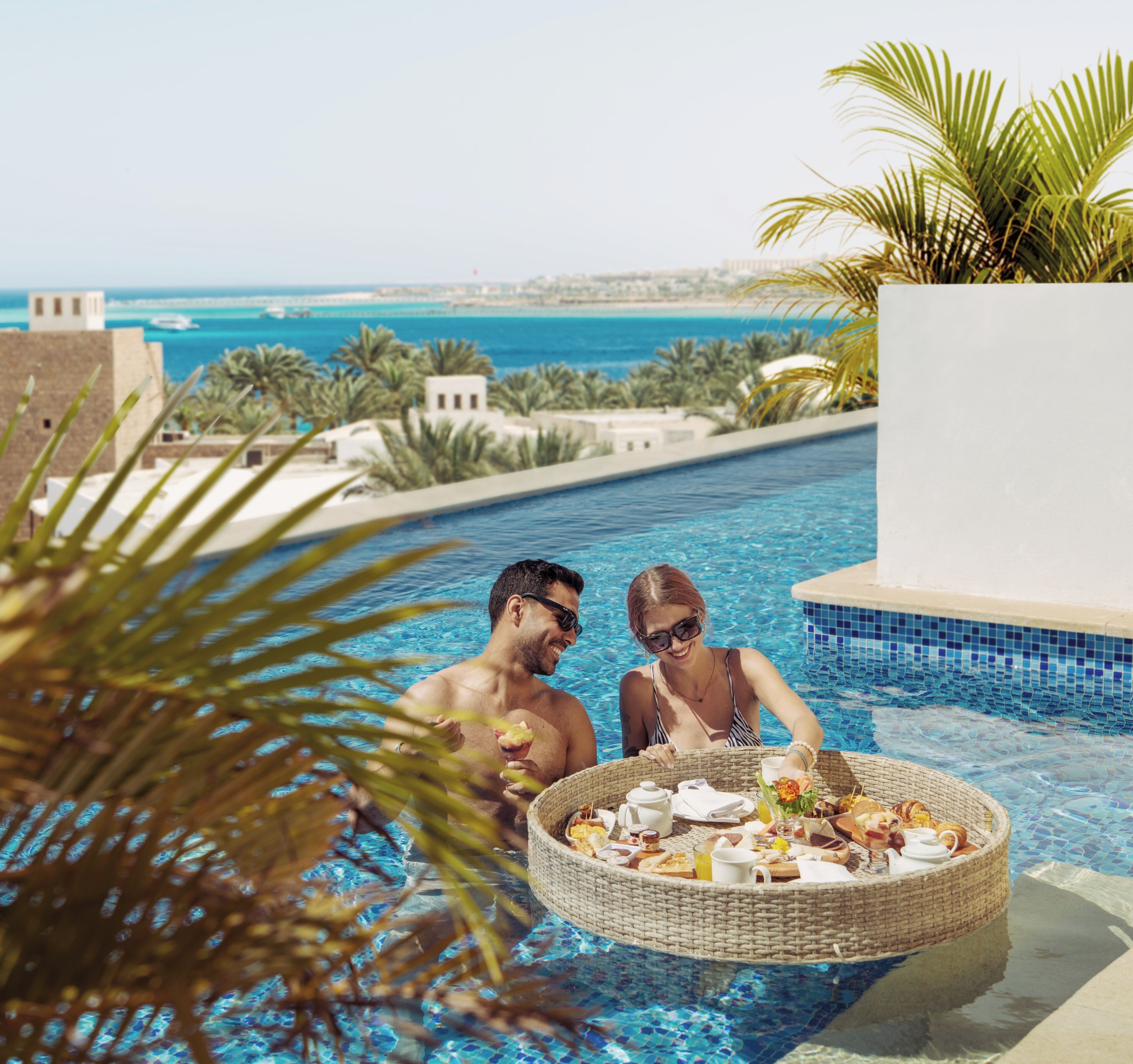 A poolside setting with a wicker basket containing food and drinks, palm trees, and a view of the sea and buildings in the background.