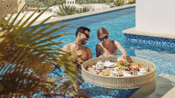A poolside setting with a wicker basket containing food and drinks, palm trees, and a view of the sea and buildings in the background.