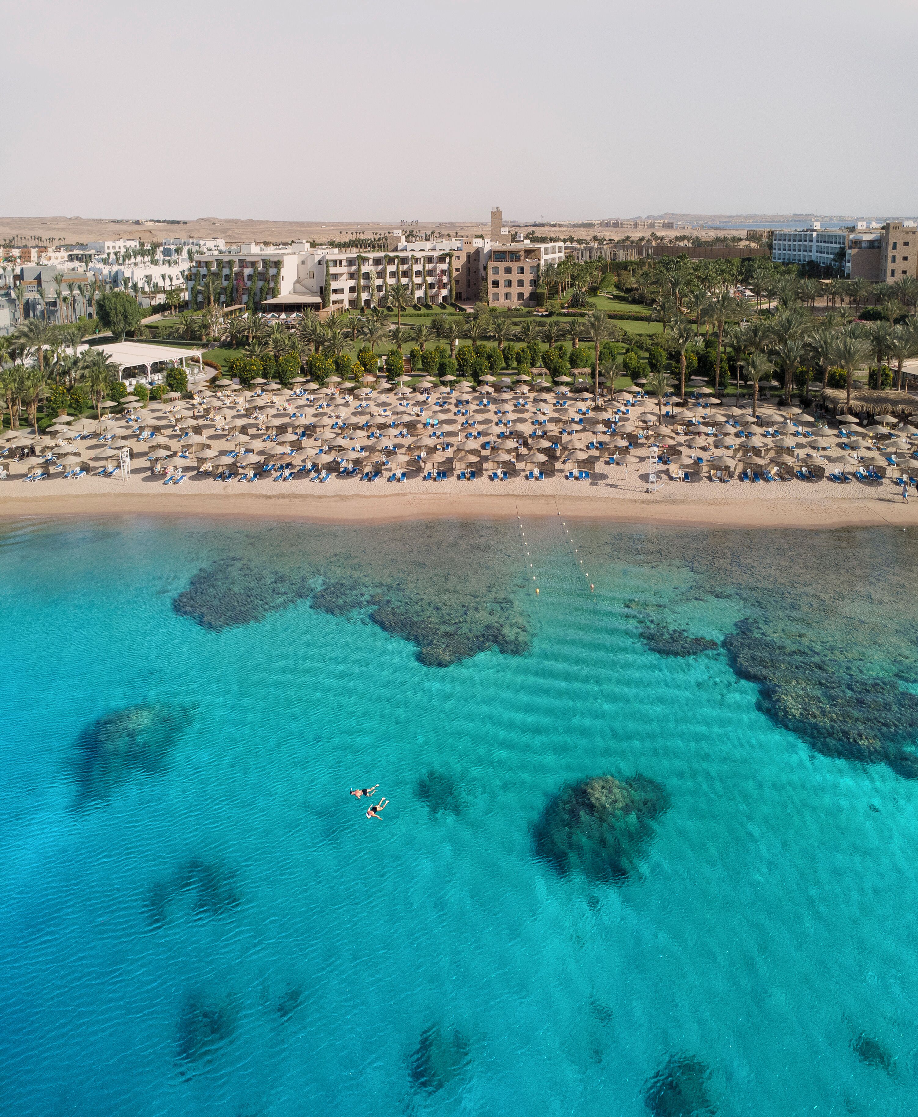 Vista aérea de un resort en la playa con aguas turquesas cristalinas, una orilla de arena y numerosos camastros y sombrillas.