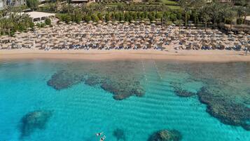 Vista aérea de un resort en la playa con aguas turquesas cristalinas, una orilla de arena y numerosos camastros y sombrillas.