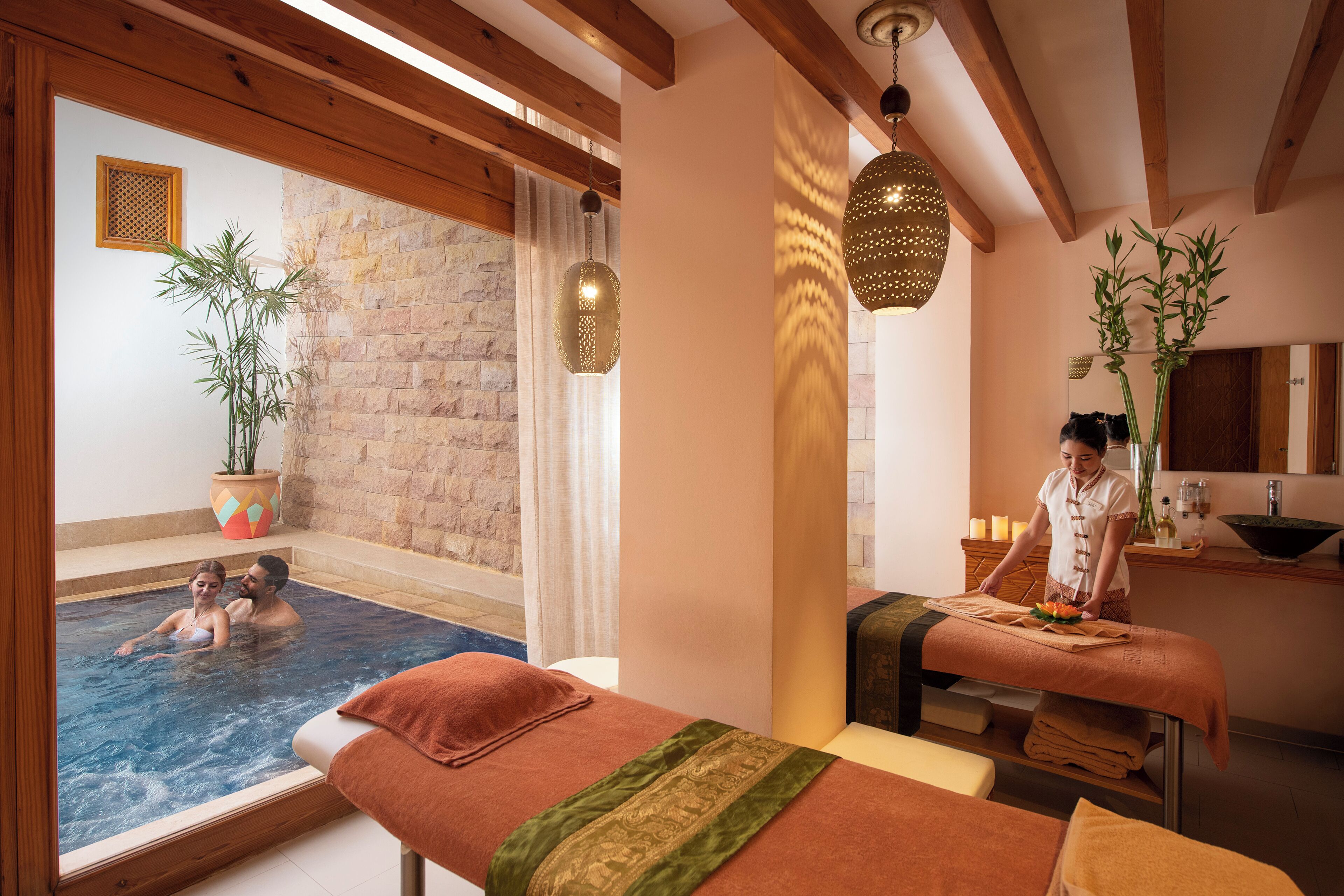 A spa room with a massage table, a massage attendant, a stone wall, and a pool area.