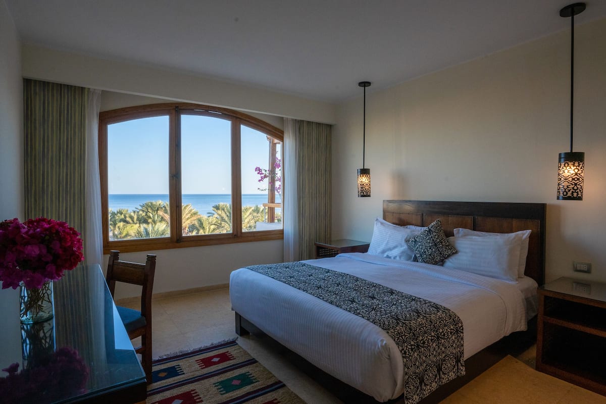 Ett hotellrum med ett stort fönster som vetter mot havet, en säng med mönstrat sänglinne, ett nattduksbord och ett skrivbord med en vas med blommor.