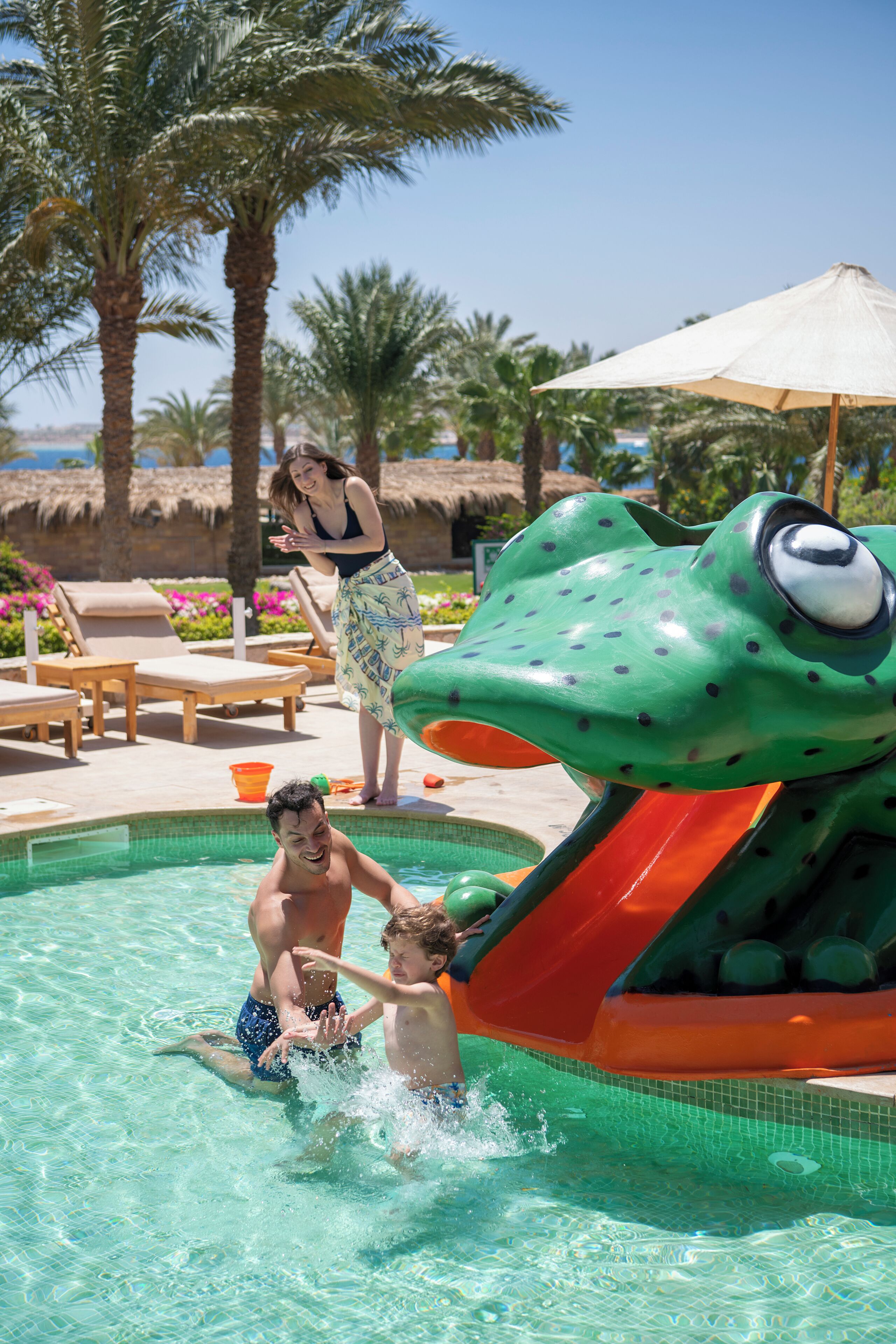 A swimming pool with a slide shaped like a frog, lounge chairs, and palm trees in the background.
