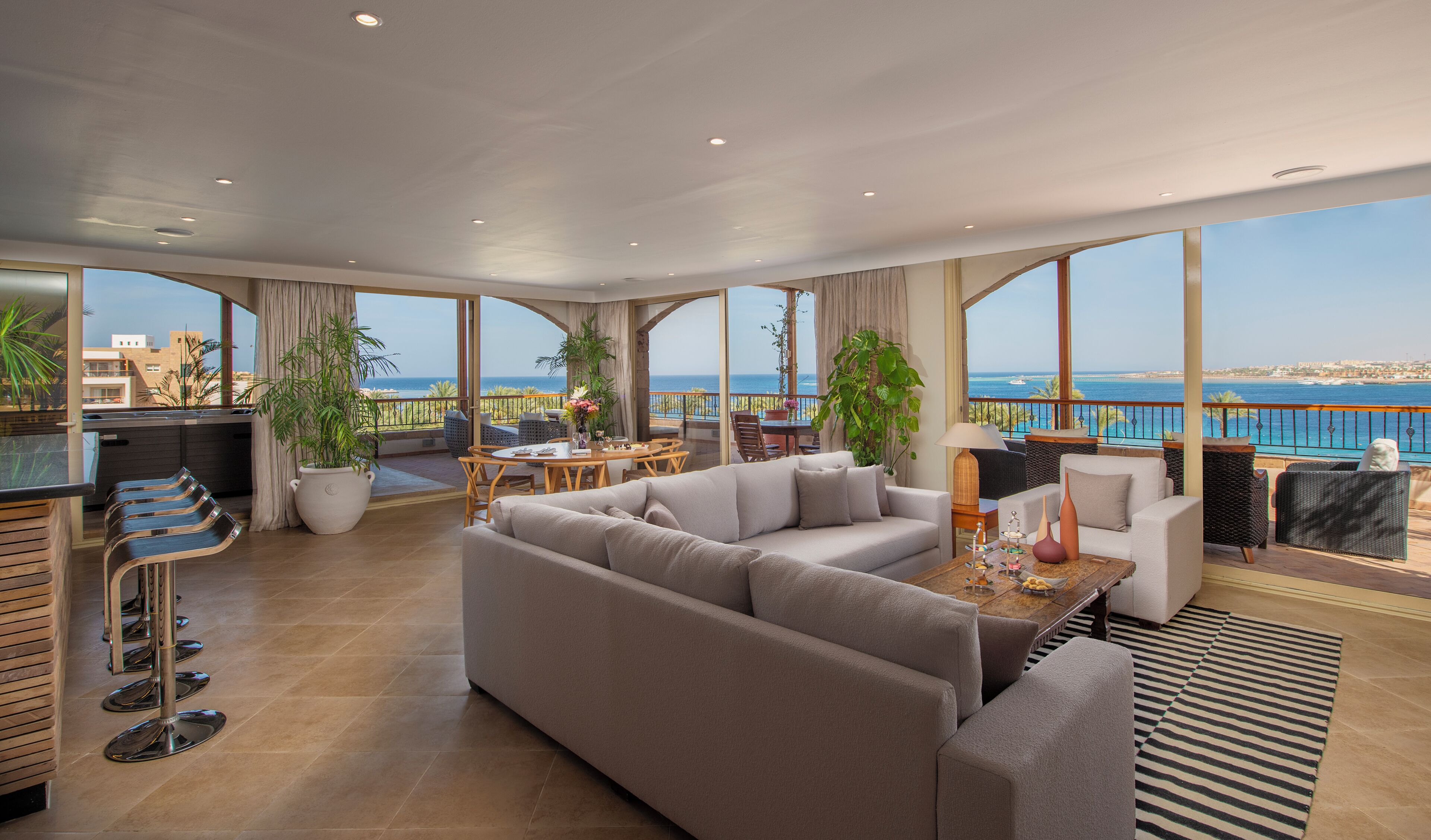 A spacious living area with a large sofa, a dining table, and a balcony with a view of the sea.