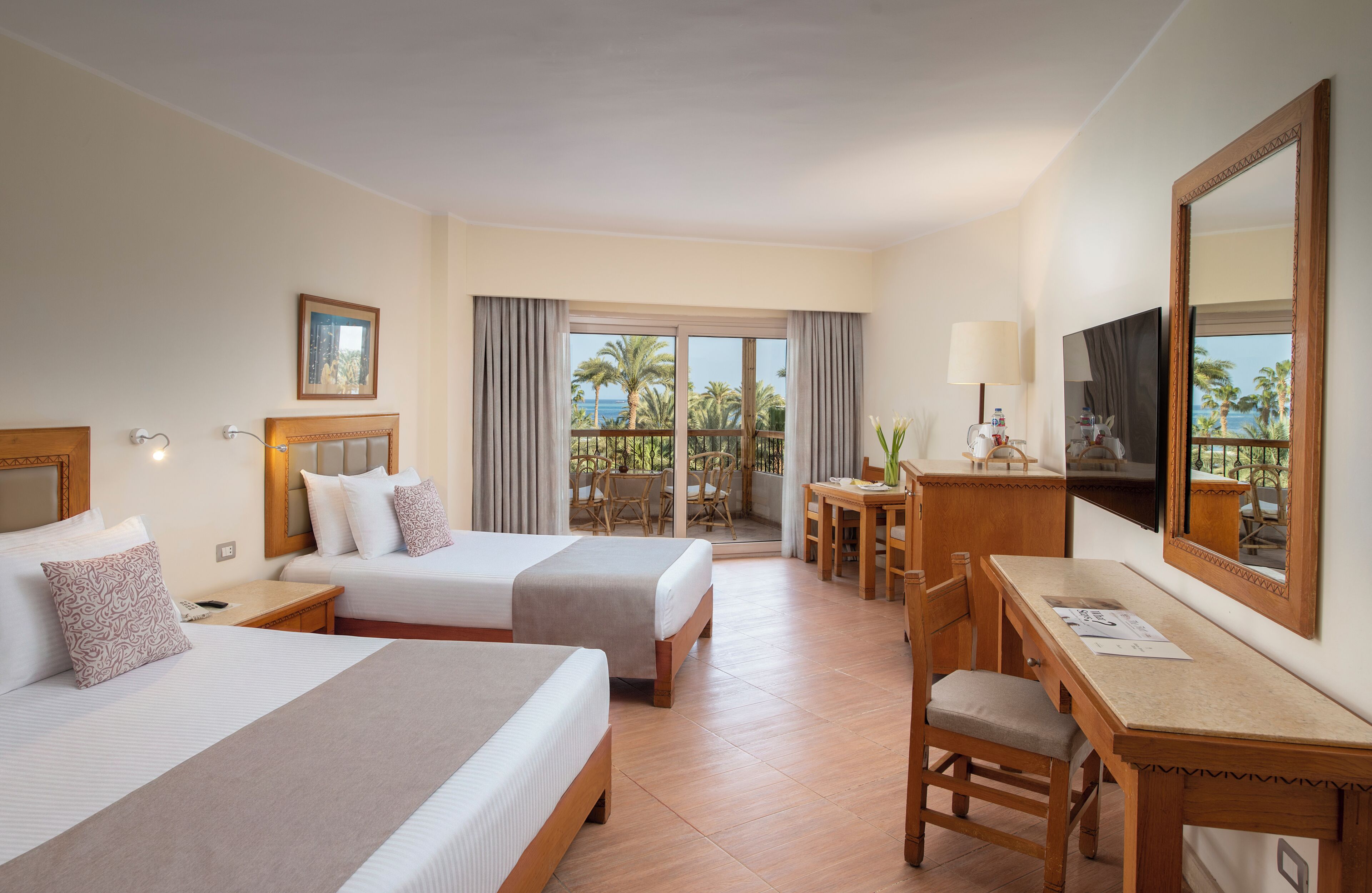 A hotel room with two beds, a desk, a chair, a TV, and a balcony with a view of palm trees and the sea.