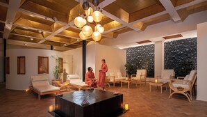 Un espace spa doté d’une plateforme centrale en pierre chauffée, d’un coin salon et d’un plafond avec des poutres apparentes et des luminaires suspendus.