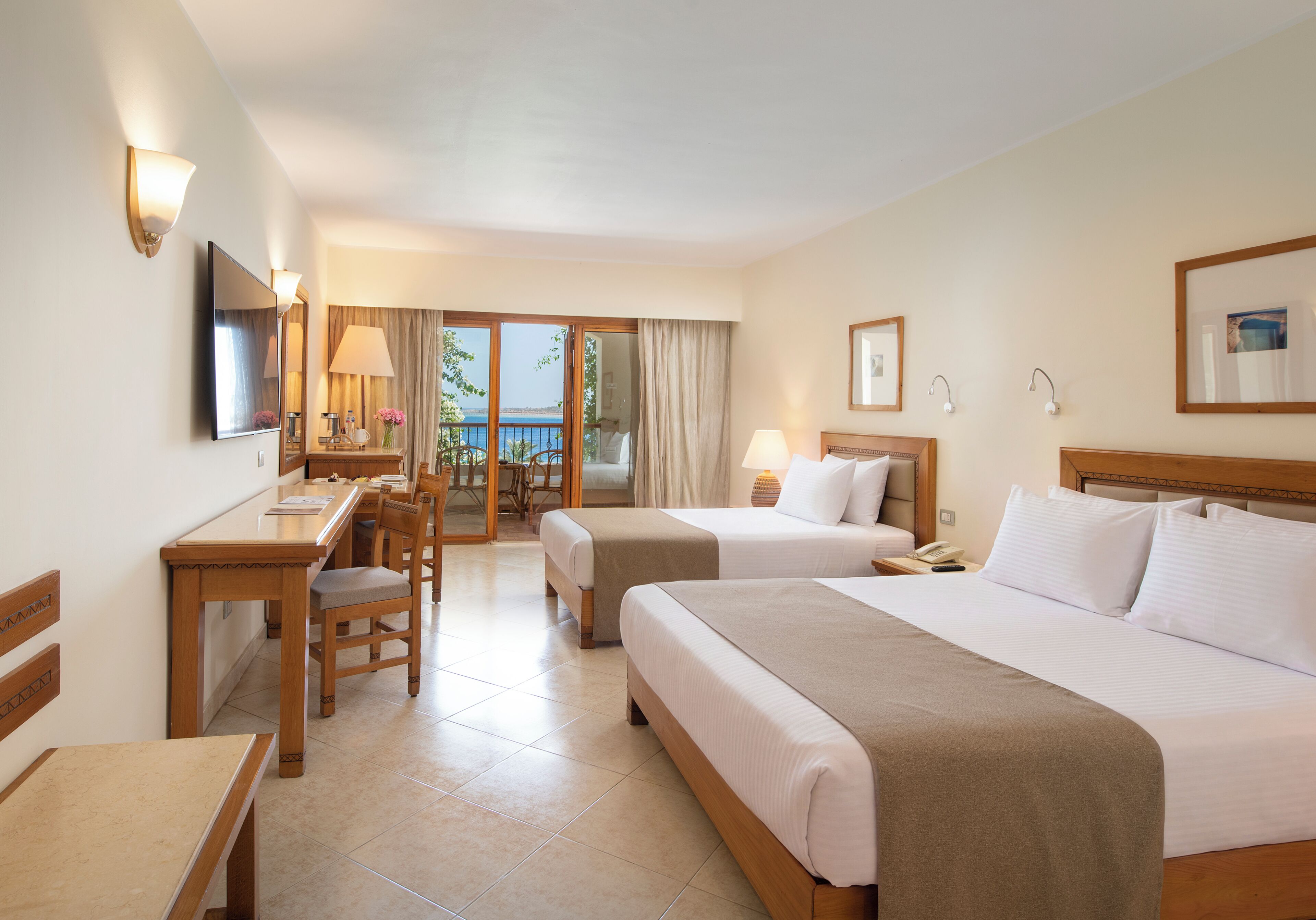 A hotel room with two beds, a desk, a chair, a TV, and a balcony with a view.