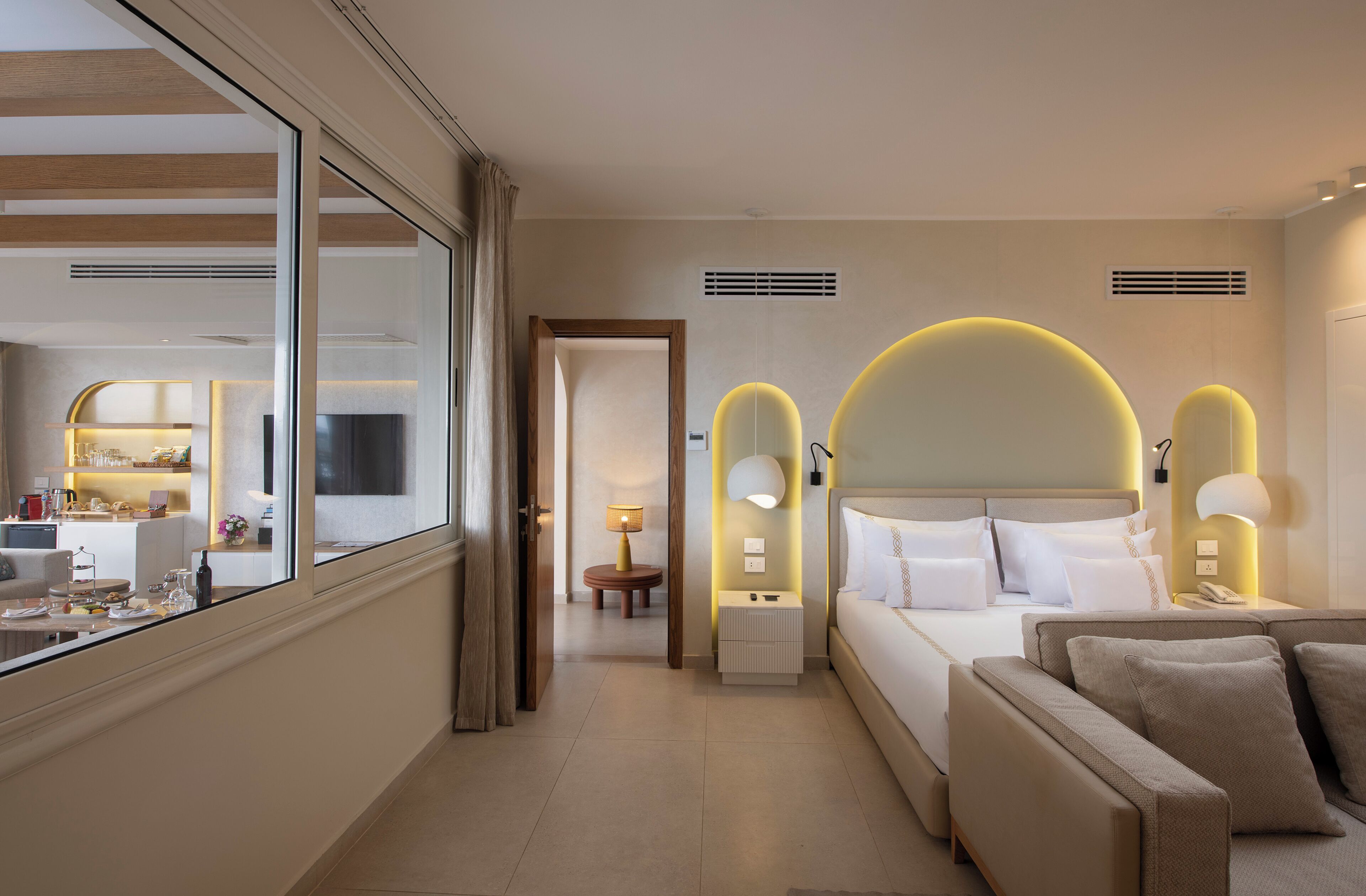 Habitación de hotel con cama, mesitas de noche, sofá, zona de comedor y vista a través de un ventanal grande.