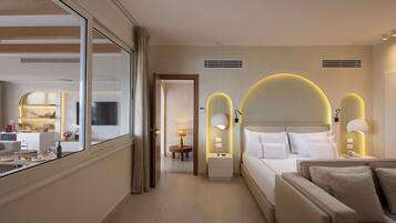 Habitación de hotel con cama, mesitas de noche, sofá, zona de comedor y vista a través de un ventanal grande.