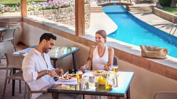 A couple dining at an outdoor table with a pool view, surrounded by wicker furniture and a stone wall.