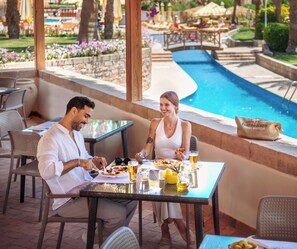 Una pareja cenando en una mesa al aire libre, con vista a una piscina, rodeada de muebles de mimbre y un muro de piedra.