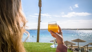 Eine Person hält ein Glas Orangensaft, im Hintergrund Palmen und der Strand.