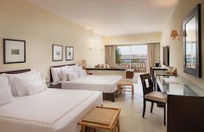 A hotel room with two beds, a desk, a chair, a small table, and a view of the sea.