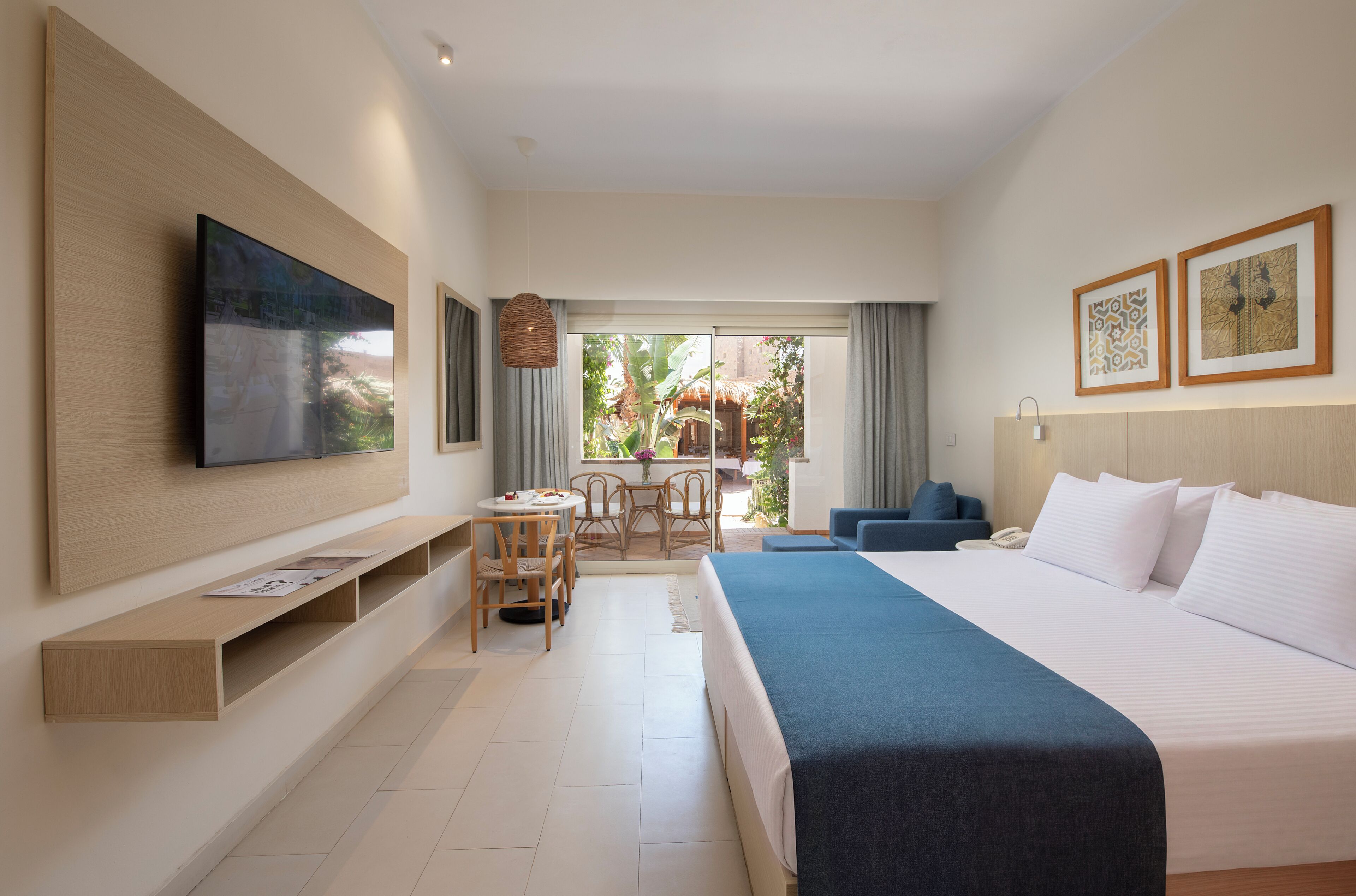 A hotel room with a large bed, a flat-screen TV mounted on the wall, a dining area with a table and chairs, and a balcony with a view of greenery.