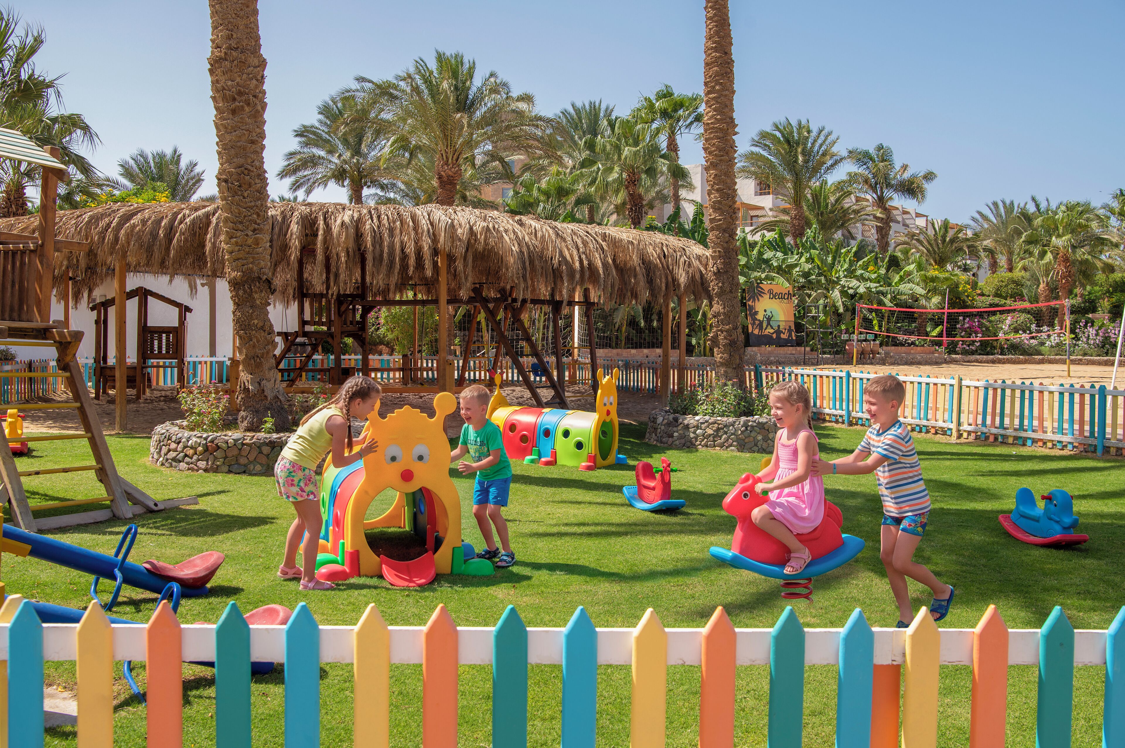 Niños jugando en un parque infantil con juegos de colores y palmeras al fondo.