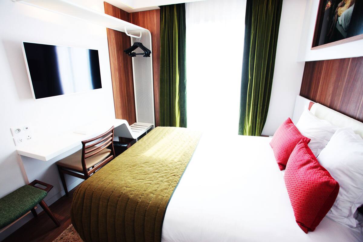 A hotel room with a bed, a desk with a laptop, a chair, and a TV mounted on the wall.