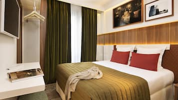 Quarto de hotel com cama, escrivaninha, cabideiro e uma janela grande com cortinas.