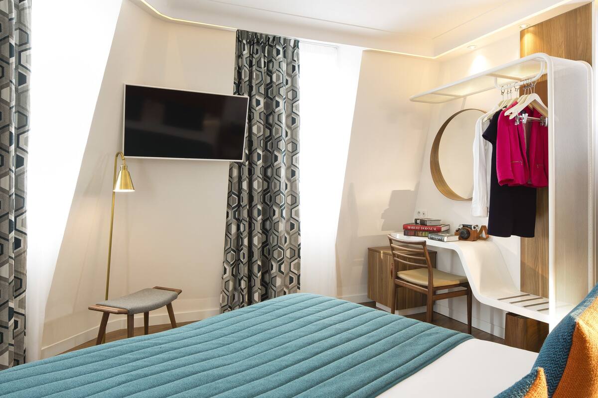 A modern hotel room with a bed, a desk, a chair, a TV, a wardrobe, and a hanging clothes rack.