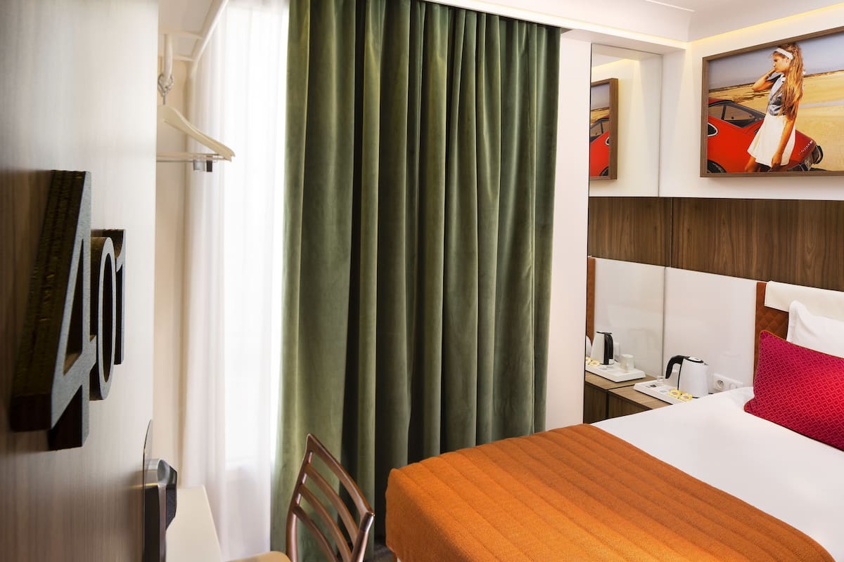 A hotel room with a bed, a desk, a chair, a window with curtains, and a painting on the wall.