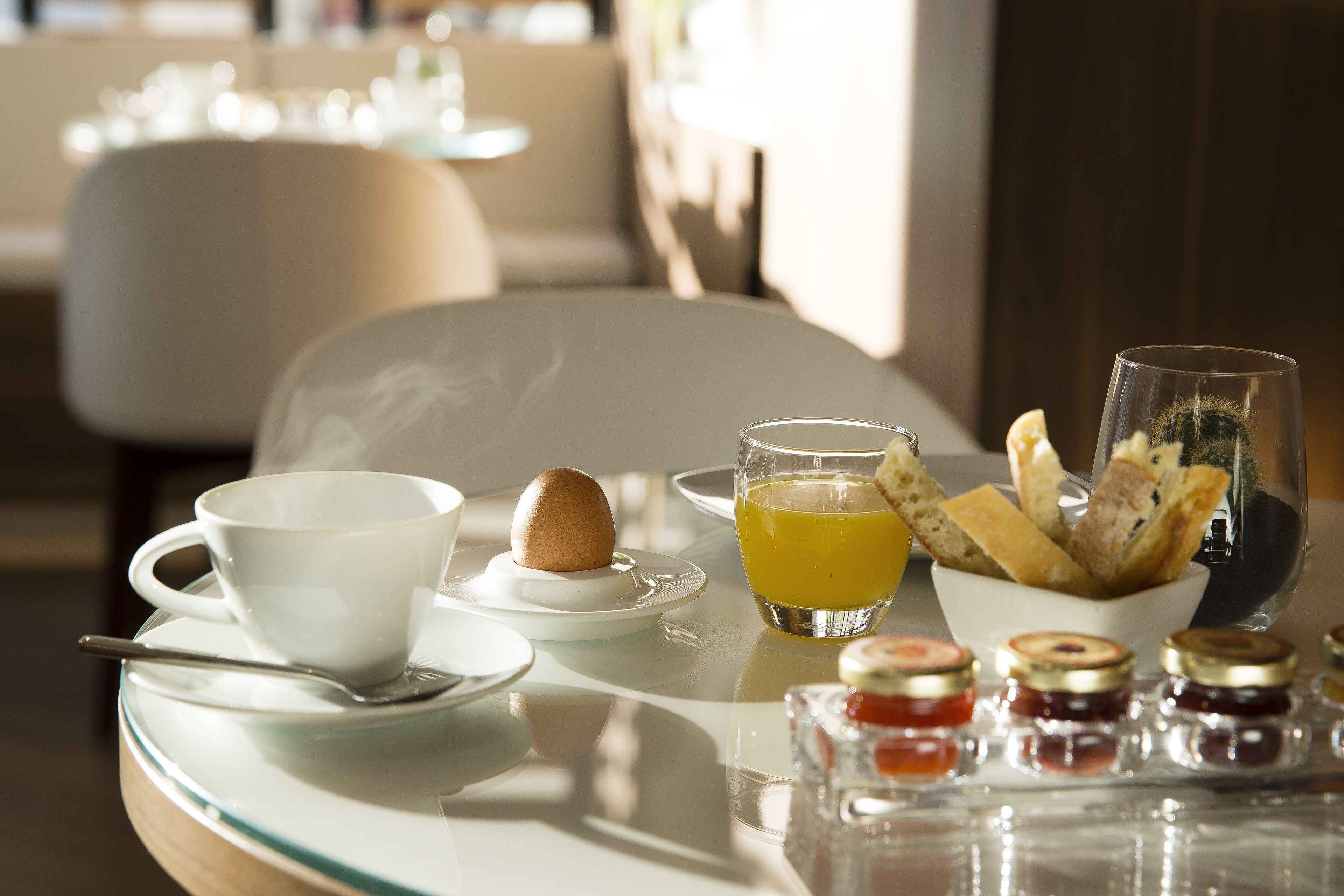 A breakfast setting with a cup, egg, orange juice, bread, and jam jars on a table.