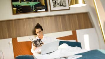 Une personne est assise sur un lit, un ordinateur portable et un livre à portée de main, dans une pièce dotée d’une tête de lit en bois, d’un tableau encadré et d’une lampe.