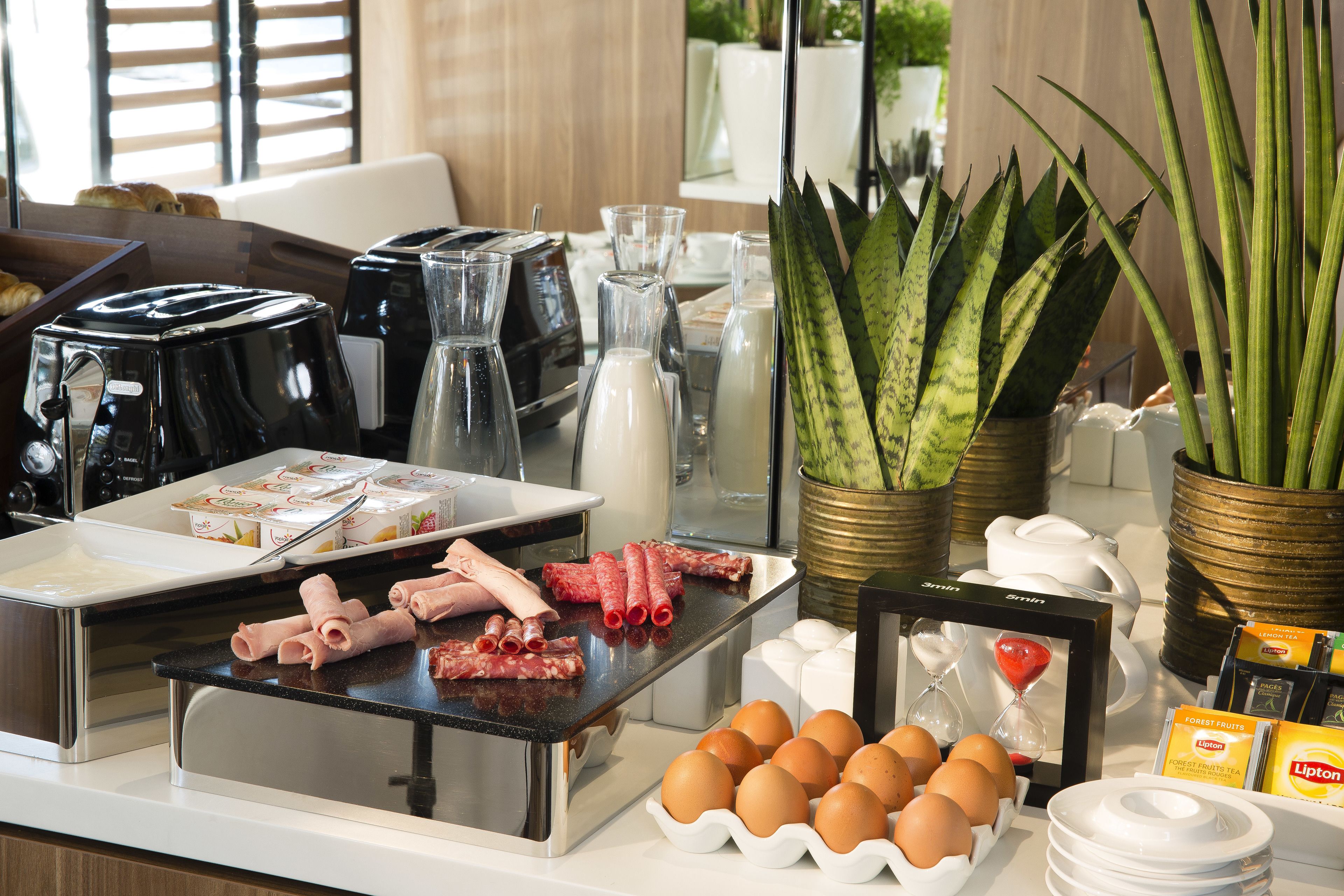 A breakfast buffet with various food items, coffee machines, and decorative plants.