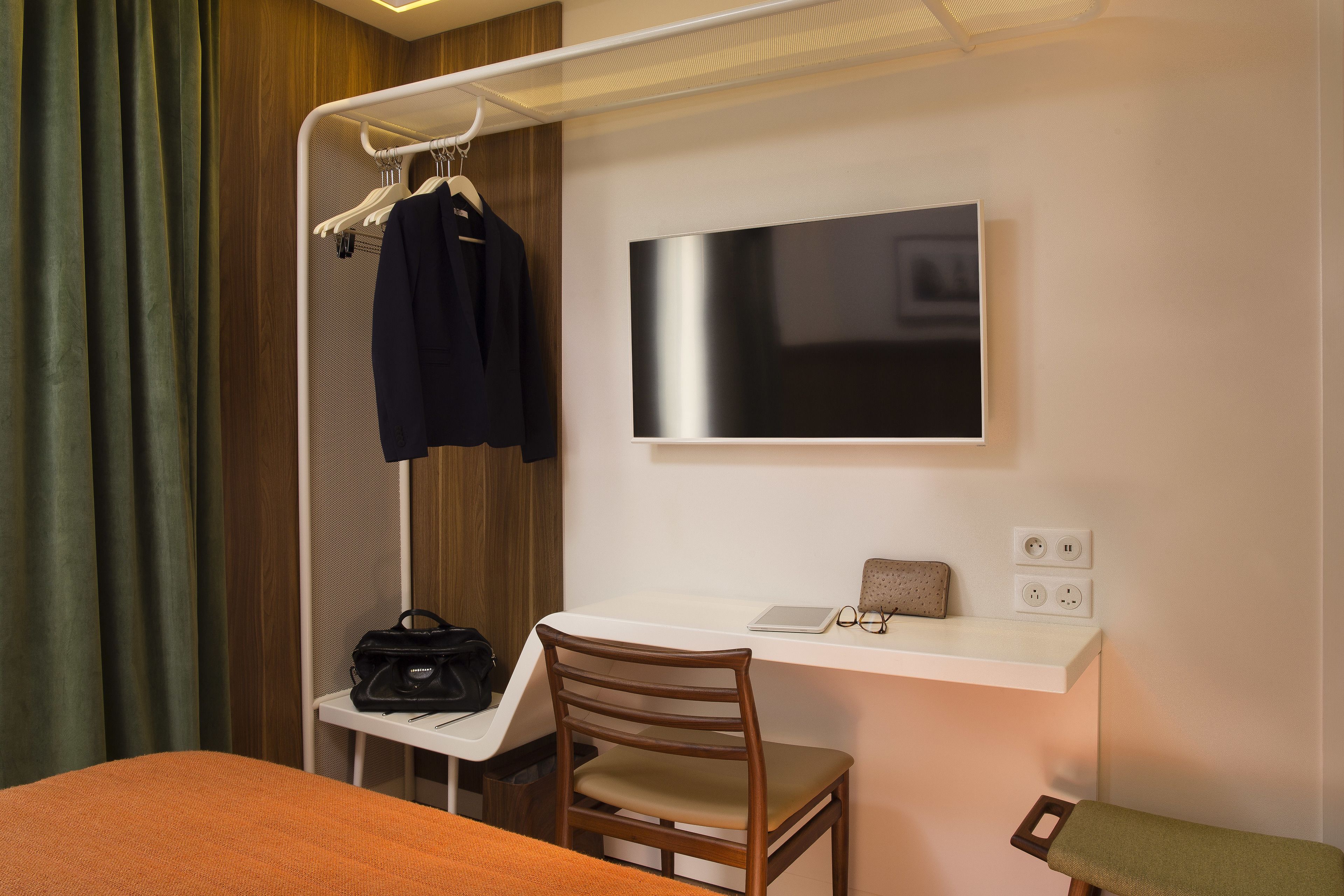 Ein kompaktes Hotelzimmer mit Bett, Schreibtisch mit Stuhl, Fernseher und Kleiderhaken mit Jacke.