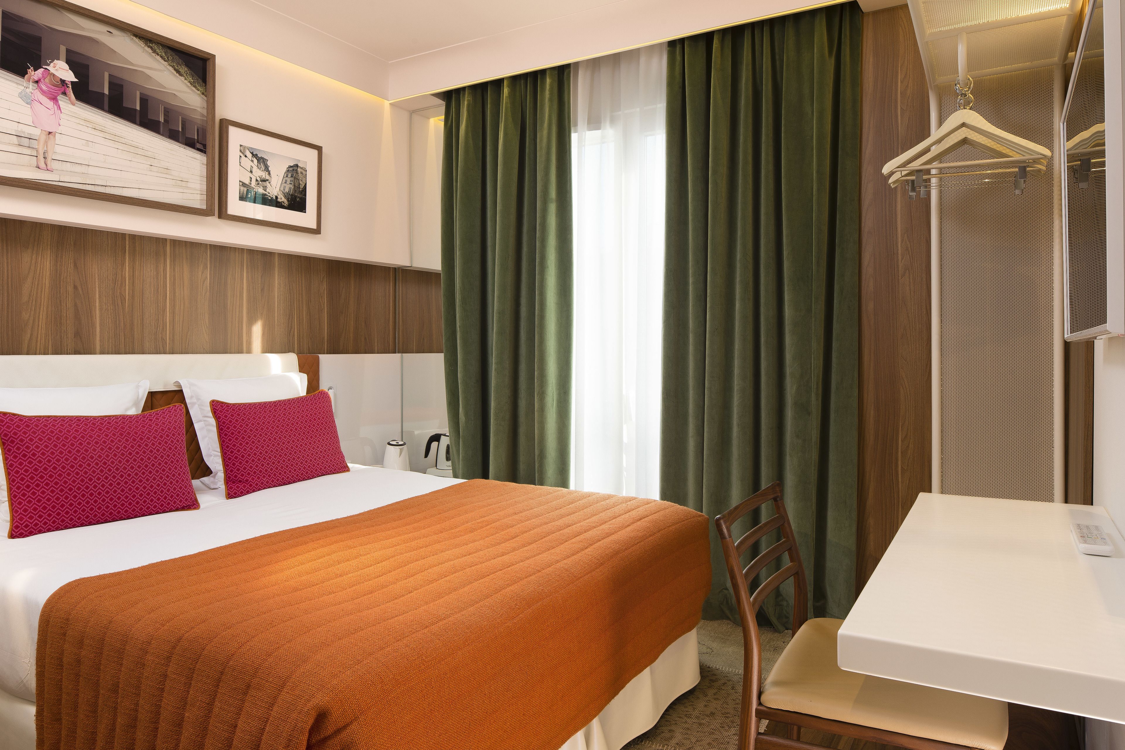 A hotel room with a bed, a desk, a chair, a hanging coat rack, and a large window with curtains.