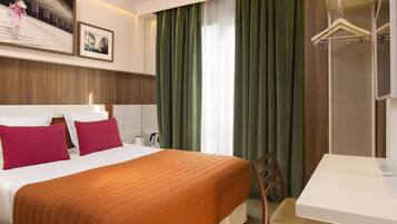 A hotel room with a bed, a desk, a chair, a hanging coat rack, and a large window with curtains.