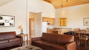 A spacious living area with a brown leather sofa, a black ottoman, and a dining table with chairs. A kitchen is visible in the background with wooden cabinets and a window with blinds. - King's Pointe Waterpark Resort (Storm Lake)