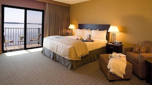 A hotel room with a large bed, two bedside lamps, a nightstand, a chair, a small table, and a view of the water through a sliding door. - King's Pointe Waterpark Resort (Storm Lake)