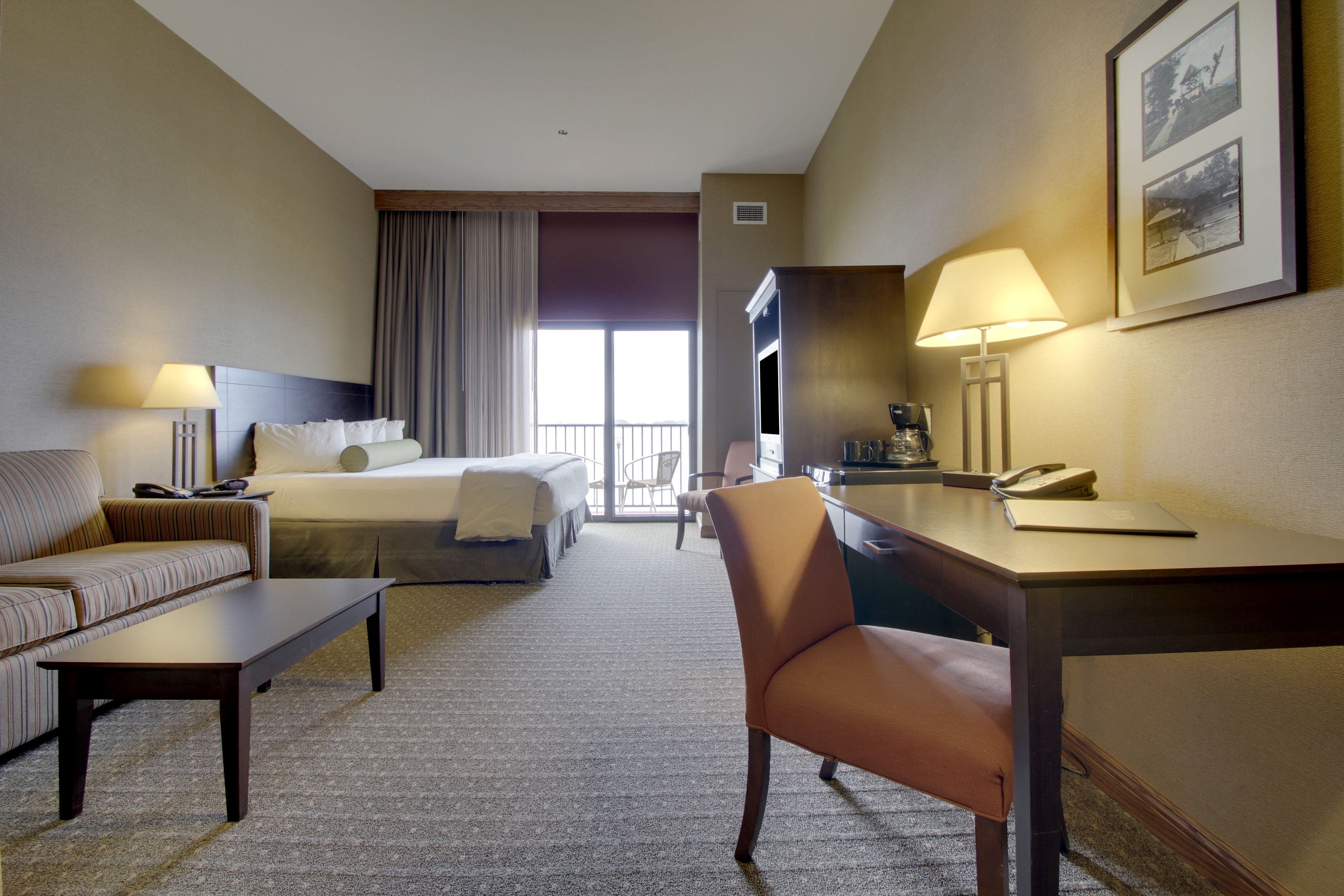 A hotel room with a bed, a sofa, a desk, and a chair. There is a window with curtains, a lamp, and a framed picture on the wall.