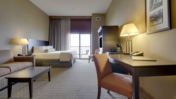 A hotel room with a bed, a sofa, a desk, and a chair. There is a window with curtains, a lamp, and a framed picture on the wall.