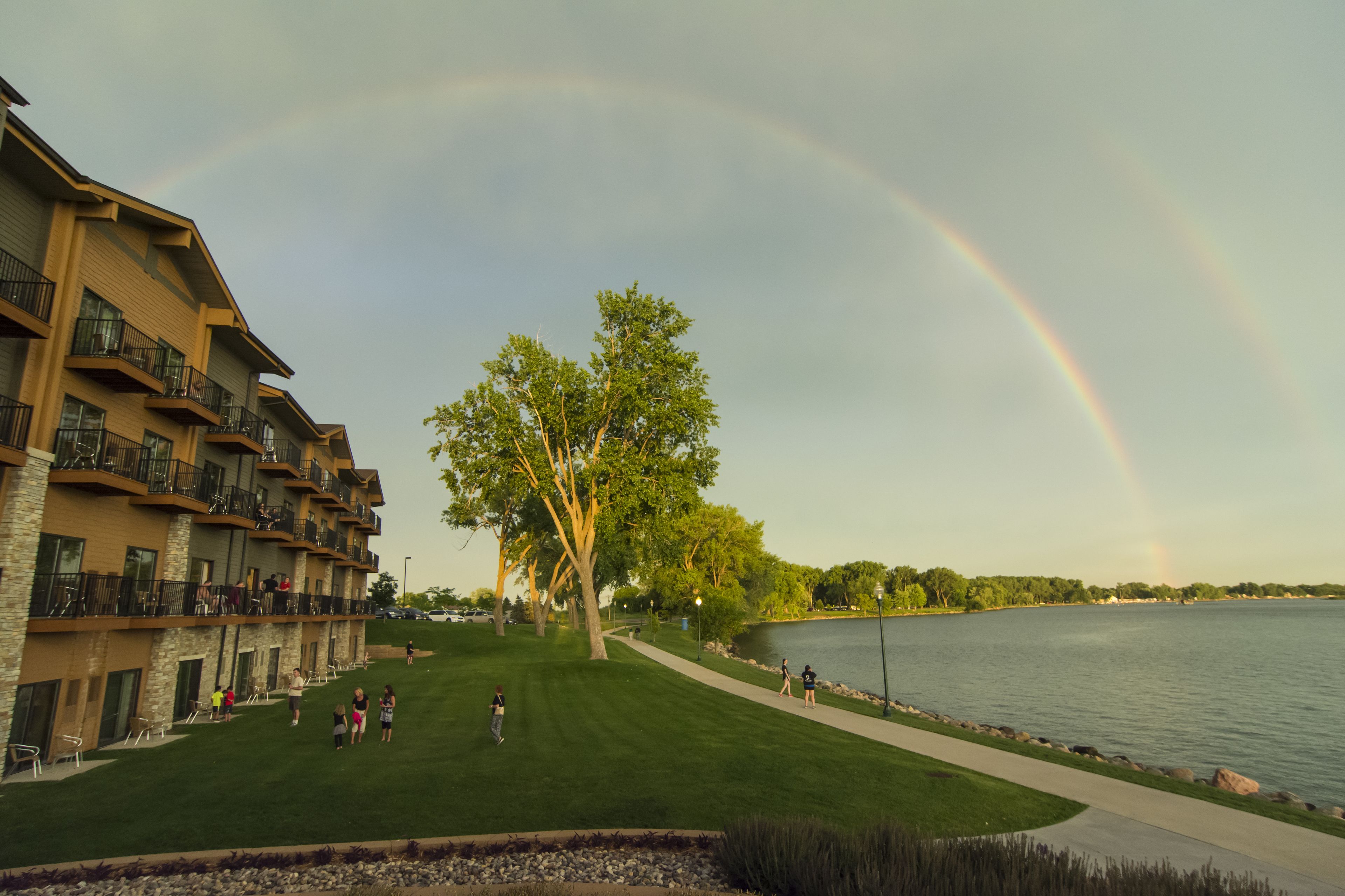 A building with multiple balconies overlooking a lake, a grassy area with people, and a visible rainbow.