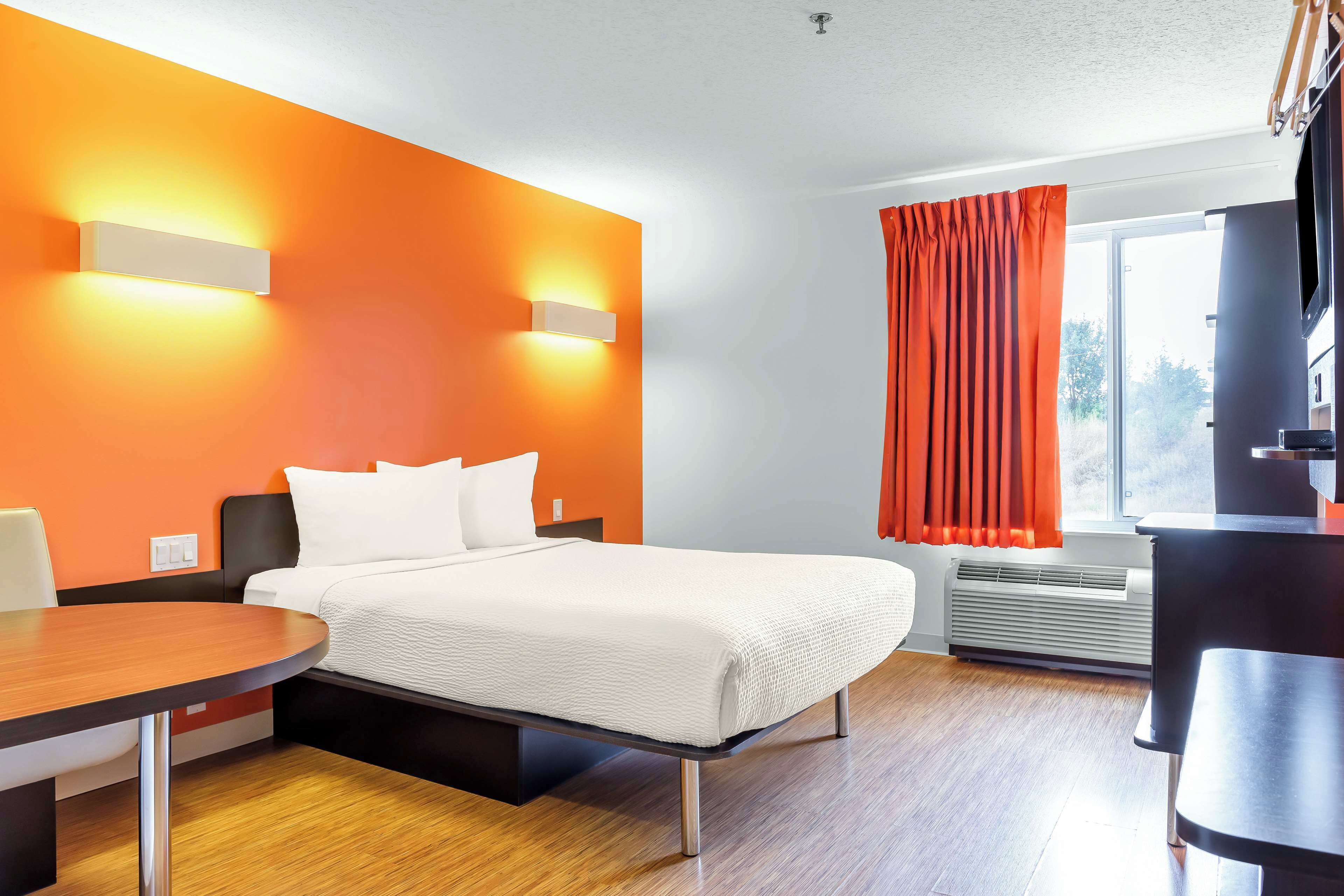 Une chambre d’hôtel avec un lit, un bureau et une chaise. Il y a une fenêtre avec des rideaux, un climatiseur et un téléviseur.