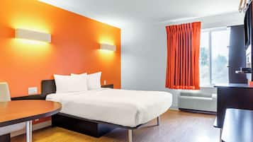 Une chambre d’hôtel avec un lit, un bureau et une chaise. Il y a une fenêtre avec des rideaux, un climatiseur et un téléviseur.