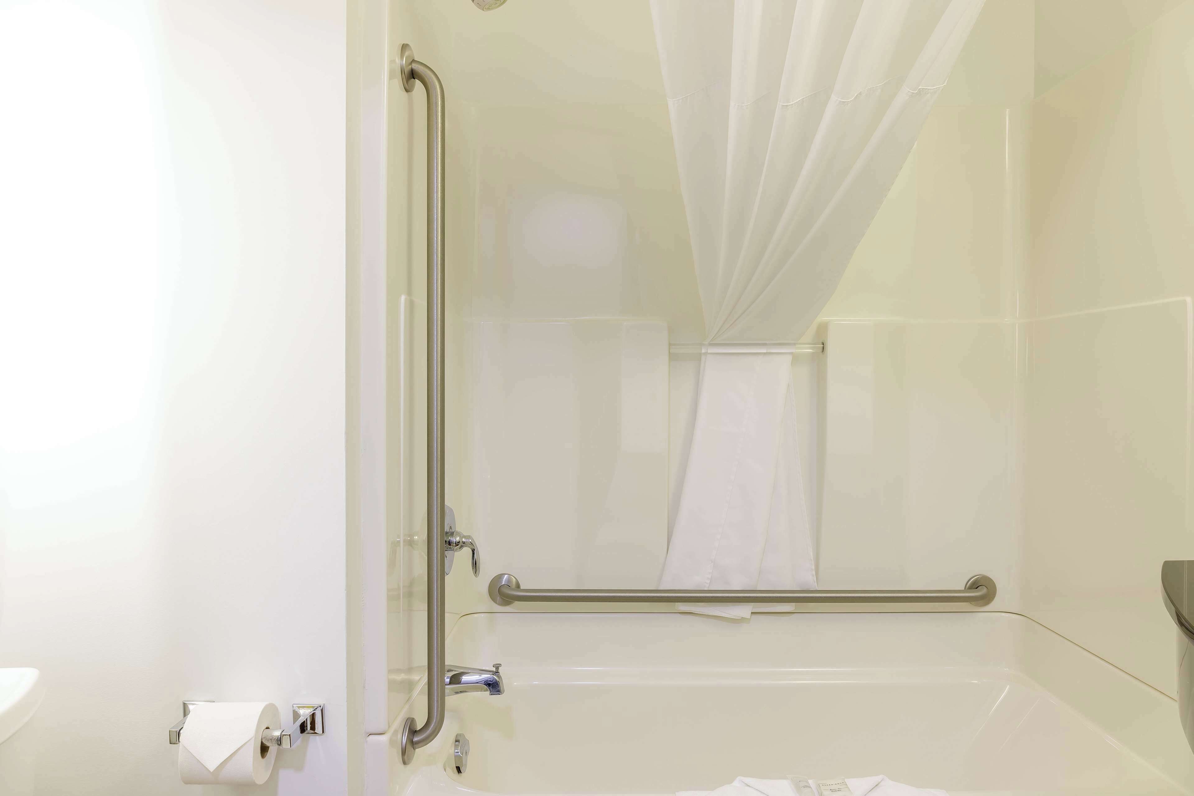 A bathroom with a glass shower enclosure, a white bathtub, a handrail, and a toilet paper holder.