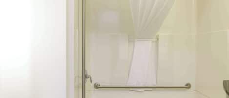 A bathroom with a glass shower enclosure, a white bathtub, a handrail, and a toilet paper holder.