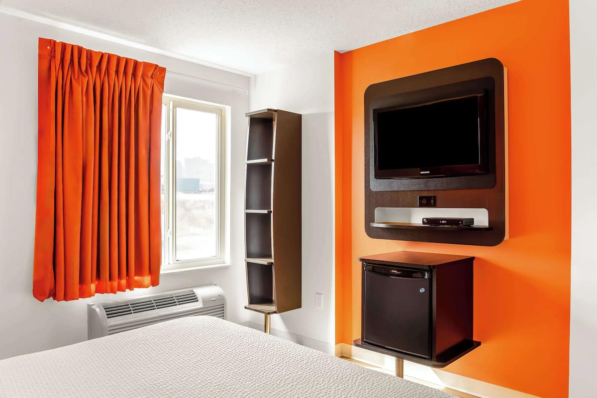Une chambre d’hôtel avec un lit, un téléviseur fixé au mur, un petit réfrigérateur et une fenêtre avec des rideaux orange.
