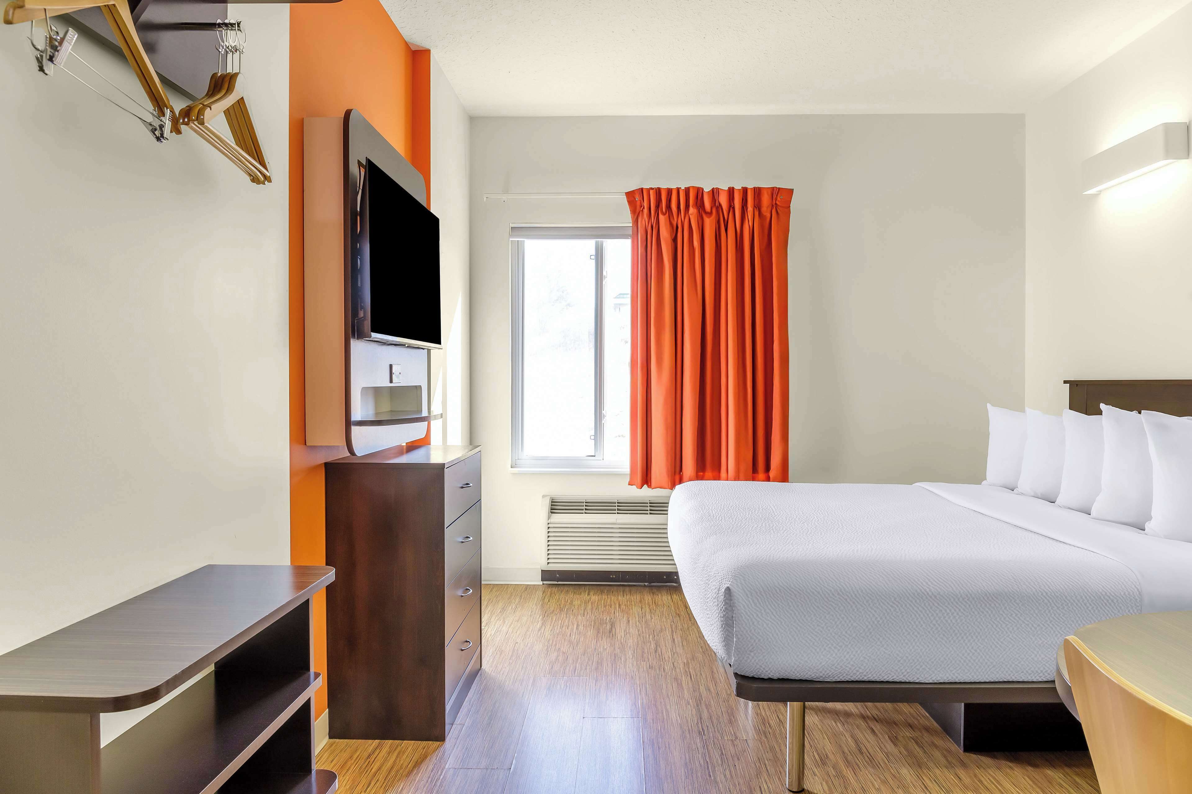 A hotel room with a bed, a TV on a wooden stand, a heater, and a window with orange curtains.