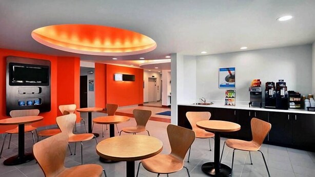 A modern cafe with round tables and orange chairs, a coffee machine, and a TV screen on the wall. - Motel 6 Swift Current, SK (Swift Current)