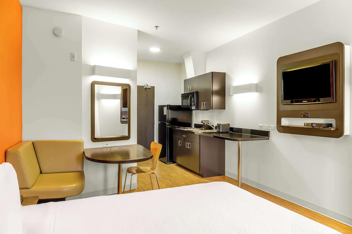 Une chambre d’hôtel comprenant un lit, un coin salon avec une chaise et une table, une kitchenette équipée d’un four à micro-ondes et un téléviseur fixé au mur.