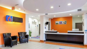 A hotel reception area with a reception desk, two black leather chairs, and a blue and red carpet with the hotel logo. - Motel 6 Swift Current, SK (Swift Current)