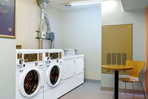 A laundry area with two front-loading washers and dryers, a small table, and a chair. - Motel 6 Swift Current, SK (Swift Current)