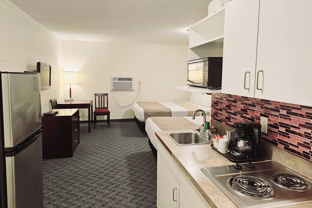 A compact kitchen with a refrigerator, microwave, stove, and sink. There is a dining table with chairs and a small desk with a lamp.