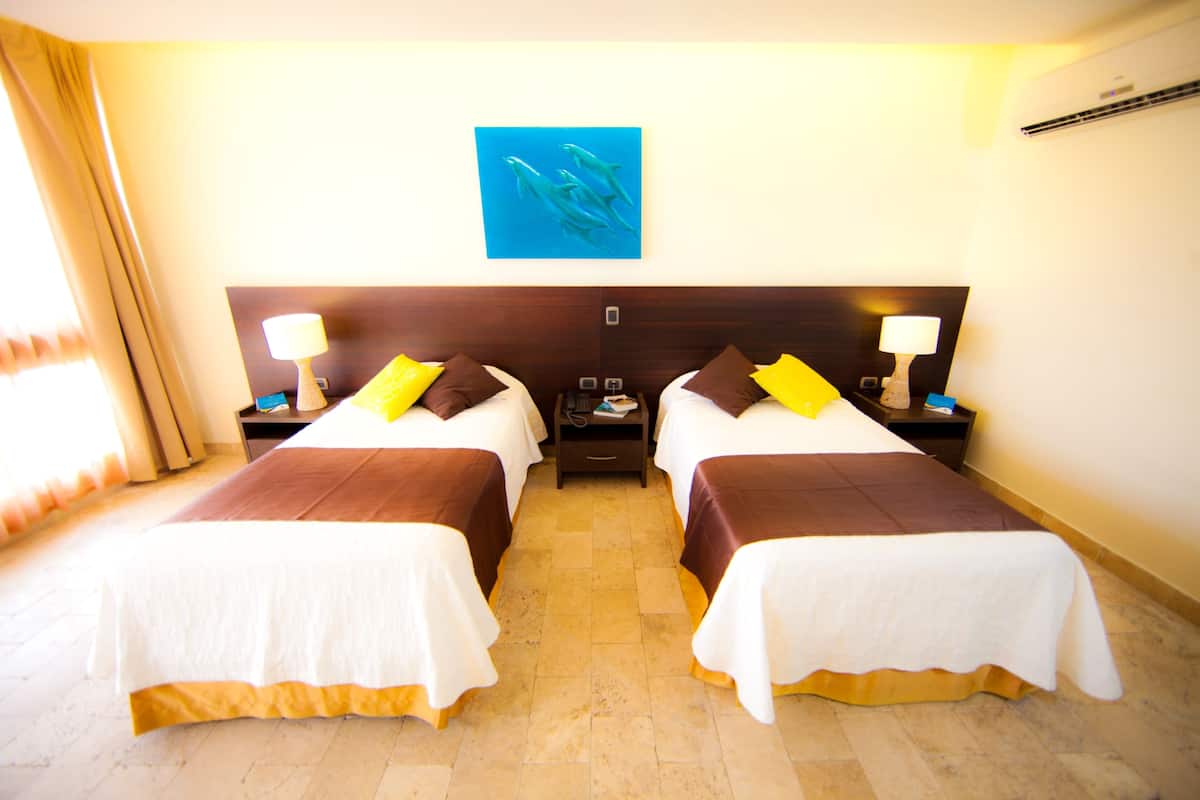 Habitación de hotel con dos camas, una mesita de noche con lámpara, un cuadro en la pared y una ventana con cortinas.