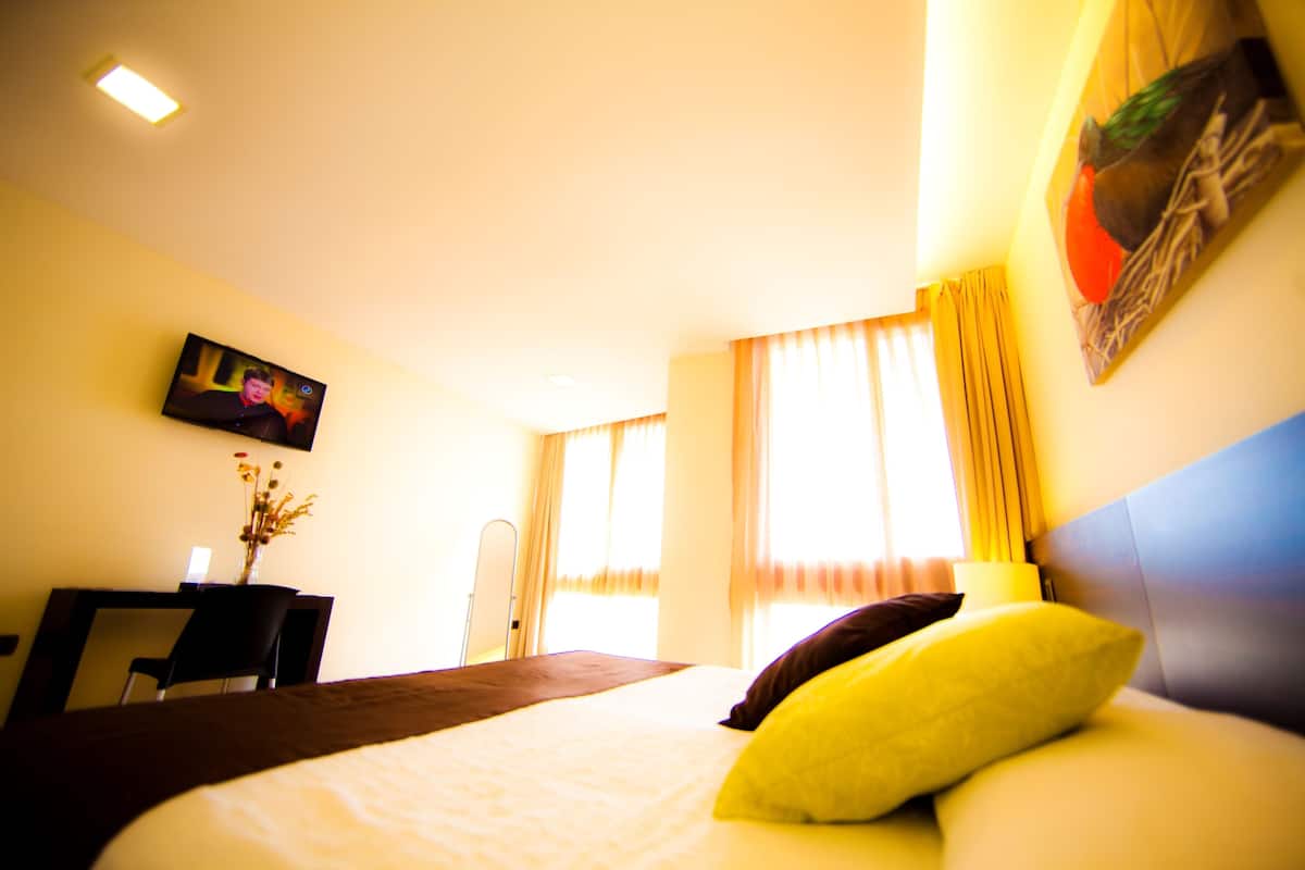Habitación de hotel con cama, televisor montado en la pared, escritorio con silla y ventana con cortinas.